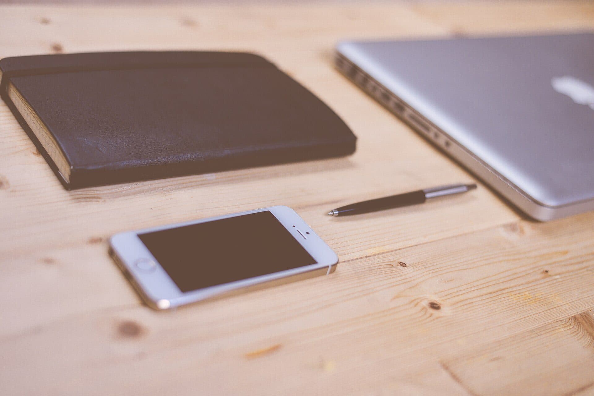A smartphone on a wooden desk beside a notebook, a pen and a laptop
