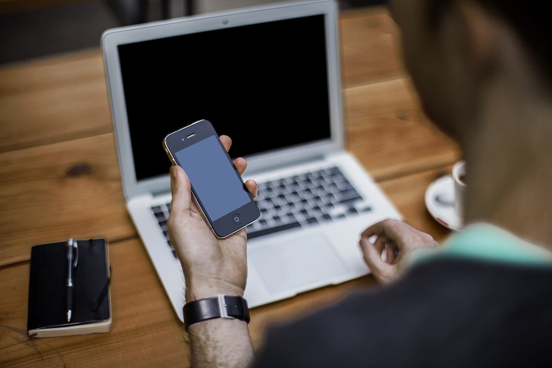 Someone at a wooden table holding a smartphone in front of a laptop, seen from behind